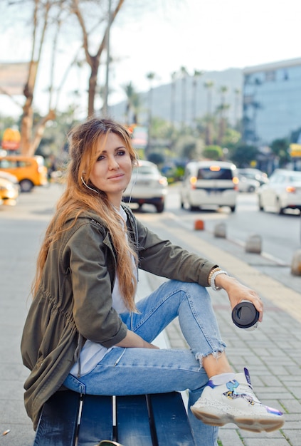 Portrait of lovely woman sitting on a bench holding coffee and listening music at street during daytime.