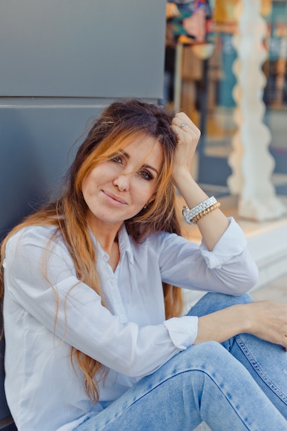 Portrait of lovely woman sitting on ground smiling at street during daytime..