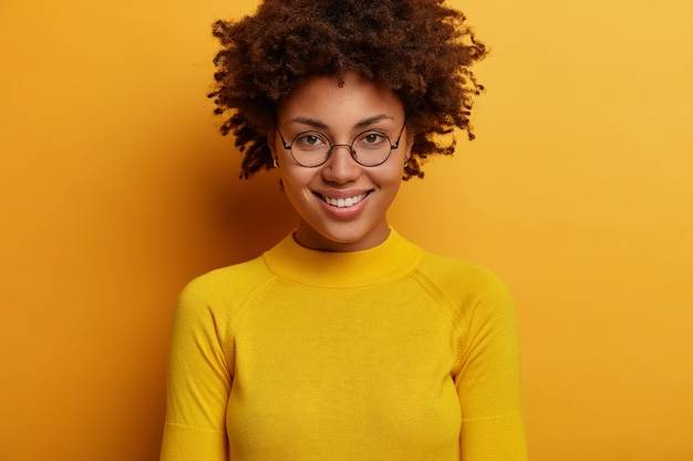 Portrait of lovely woman smiles gently, has curly hair, wears round transparent glasses and yellow jumper, looks directly , listens pleasant news, poses indoor. human face expressions