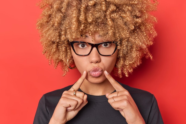 Portrait of lovely woman with curly blonde hair keeps lips folded indicates index fingers at mouth wears transparent glasses and casual black t shirt poses against red background sends air kiss