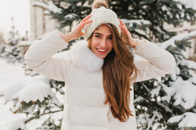 Portrait of  lovely woman with long light-brown hair showing true happy emotions in winter day on fir tree. Charming young woman in white jacket fooling around in cold morning at snowy park.