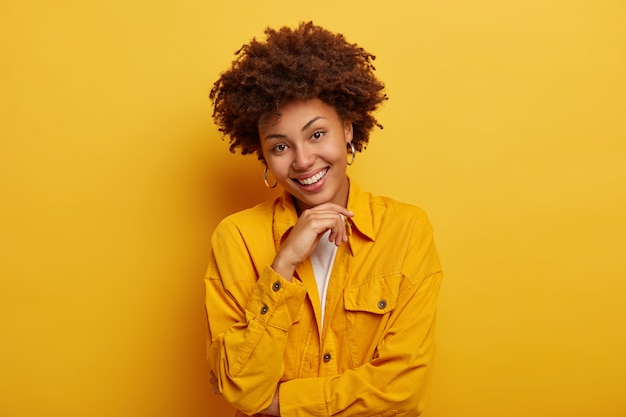 Free photo portrait of lovely woman with natural beauty, afro hairstyle, wears bright yellow jacket, big earrings, touches chin, has carefree expression, poses indoor at studio.