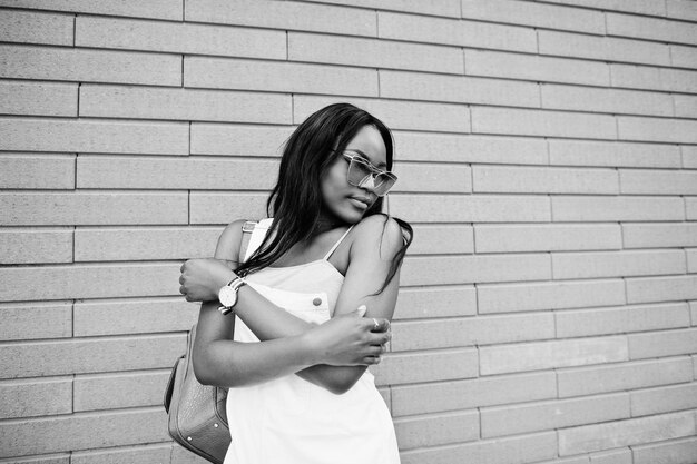 Portrait of a lovely young african american woman posing with sunglasses against a brick wall in the background Black and white photo