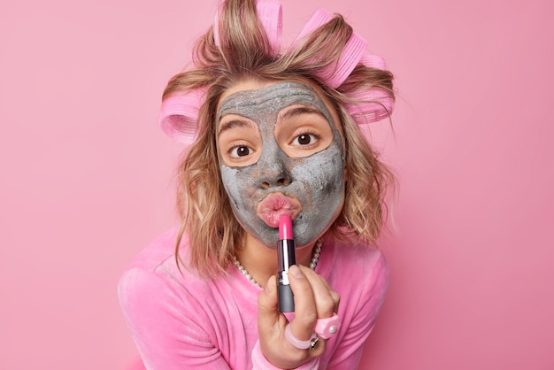 素敵な若いヨーロッパの女性の肖像画は、口紅の美しさを養うマスクを適用し、ヘアローラーは、ピンクの背景の上に分離された化粧品を使用して美しく見えたい日付の準備をします