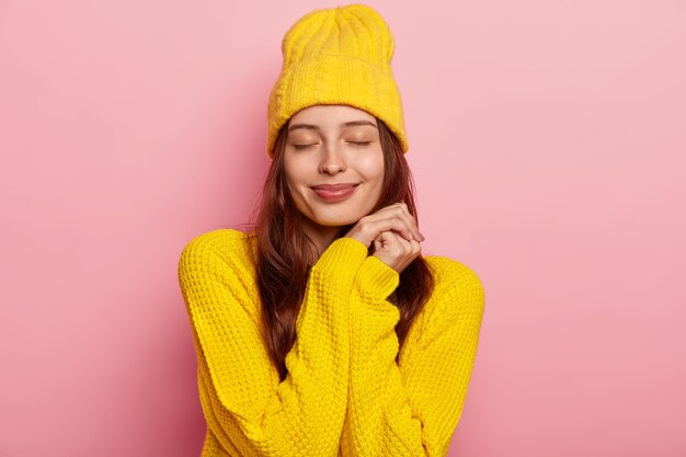 Portrait of lovely young European woman keeps eyes shut, wears vivid yellow knitted sweater and headgear, isolated over pink background.