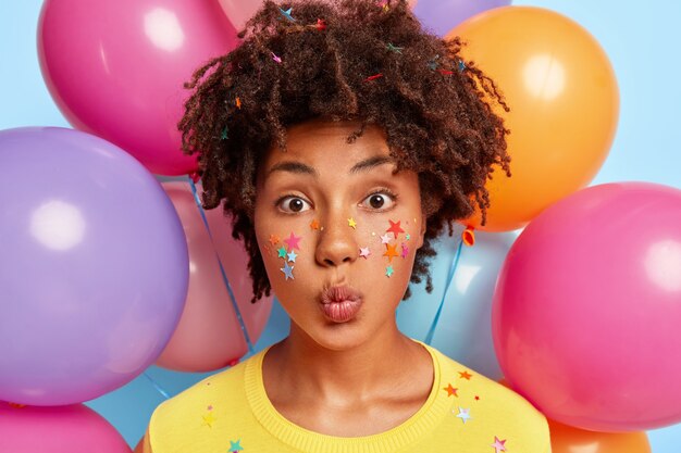 Portrait of lovely young female model with curly hairstyle, keeps lips folded, stick colorful stars on face, wears yellow clothes, makes grimace, bunch of helium airballoons in wall