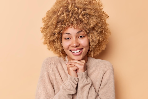 Portrait of lovely young woman keeps hands under chin looks delighted at camera feels glad smiles toothily has happy facial expression dressed in casual jumper isolated over brown background