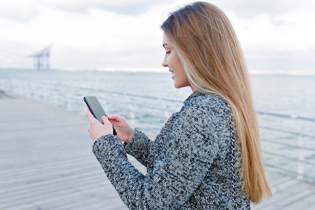 Портрет прекрасной молодой женщины со счастливой улыбкой в сером пальто, прокручивая смартфон на берегу моря