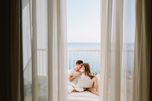 Portrait of loving couple watching movie together on balcony in morning with curtains on foreground. Young man resting with his amazing tanned girlfriend in bed holding laptop with sea views