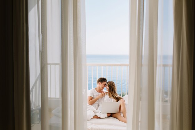 Портрет влюбленной пары вместе смотреть фильм на балконе утром с занавесками на переднем плане. Молодой человек отдыхает со своей удивительной загорелой подругой в постели с ноутбуком и видом на море
