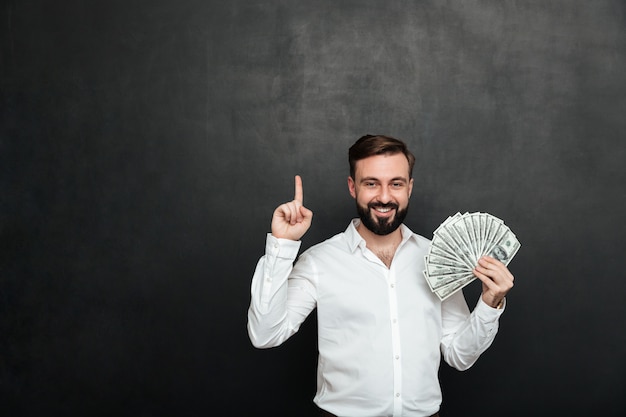 Portrait of lucky man in white shirt holding lots of money cash in hand and showing finger up over dark gray