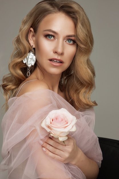 Портрет роскошной блондинки с идеальным макияжем, позирующей с розовой розой