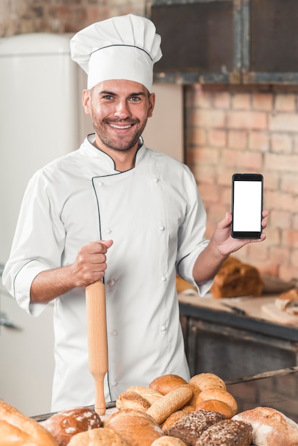 Foto gratuita ritratto del panettiere maschio che mostra cellulare in bianco con i pani al forno