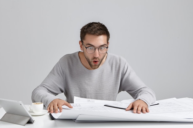 Portrait of male engineer stares at drawings, looks in surprised expression, tries to understand what is written