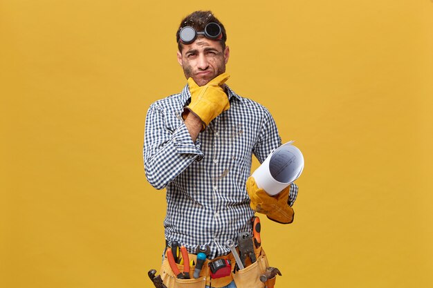 Portrait of male fettler wearing protective wear holding blueprint having thoughtful expression while thinking over his actions during work. Repairman with kit of instruments isolated over wall