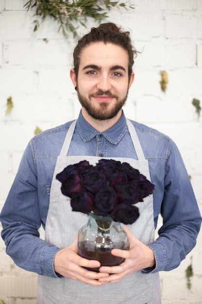 Foto gratuita ritratto di un fiorista maschio che tiene i fiori di rosa nel vaso di vetro