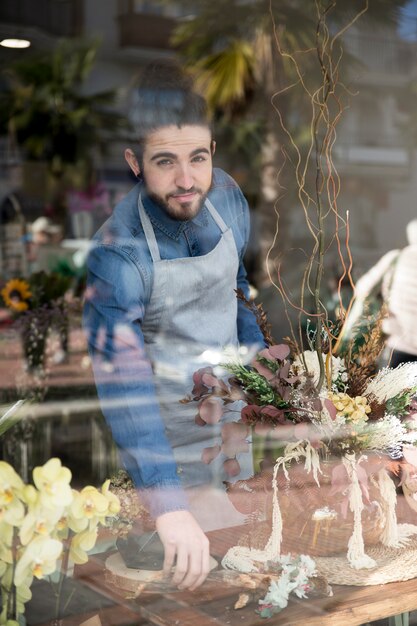 Portrait of a male florist standing behind the glass arranging the flower in his shop