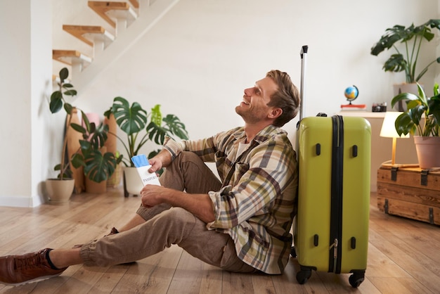 Portrait of male model with suitcase sitting on floor holding flight tickets and passport going