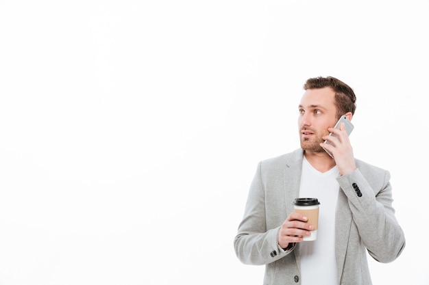 Портрет мужчины офисного работника, пить кофе на вынос, имея приятный мобильный разговор на мобильный телефон и глядя в сторону, на белую стену