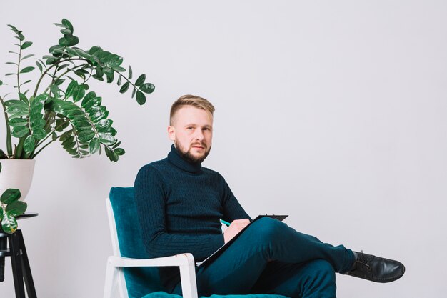 Portrait of a male psychologist sitting on chair with clipboard and pen against white wall