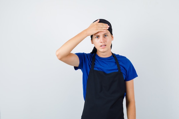 Portrait of male teen cook keeping hand on forehead in t-shirt, apron and looking stressed front view