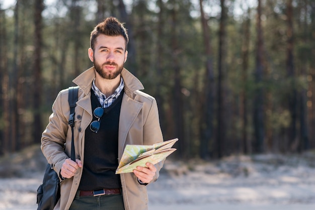 Free photo portrait of a male traveler with his backpack on shoulder holding map in hand looking at camera