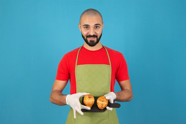 Portrait of a man in apron and gloves holding fresh pastries . 