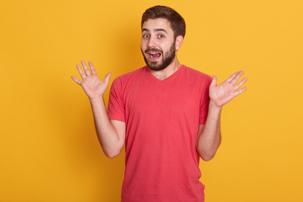 Portrait of man being surprised, handsome male spreading his hands up, posing isolated over yellow wall, attractive unshaven guy wearing red casual t shirt. The concept of human emotions.