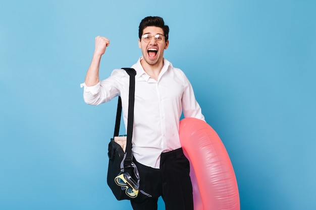 Portrait of man in business outfit enjoying start of vacation. Guy posing with inflatable circle against blue space.