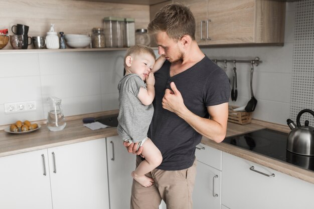 Portrait of a man carrying his little son standing in the kitchen gesturing