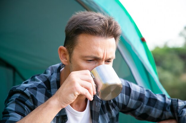 Portrait of man drinking tea, sitting in tent and thinking. Caucasian attractive male tourist sitting alone and holding metal cup.  Tourism, adventure and summer vacation concept