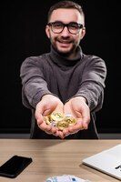 Free photo portrait of man in glasses showing golden bitcoins in his hands at desk isolated over black