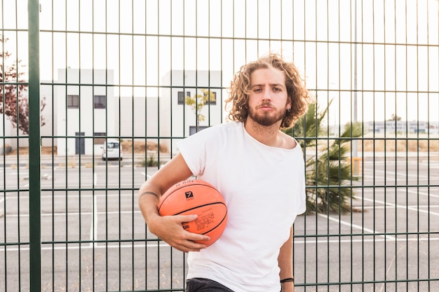 Foto gratuita ritratto di una pallacanestro della tenuta dell'uomo che sta contro il recinto