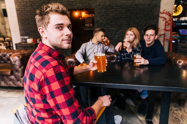 Портрет мужчины, держа бокал пива, сидя с друзьями, глядя на камеру