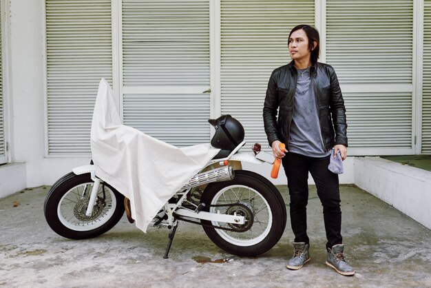 Portrait of man in leather jacket standing with polish spreay at his bike covered by throwover