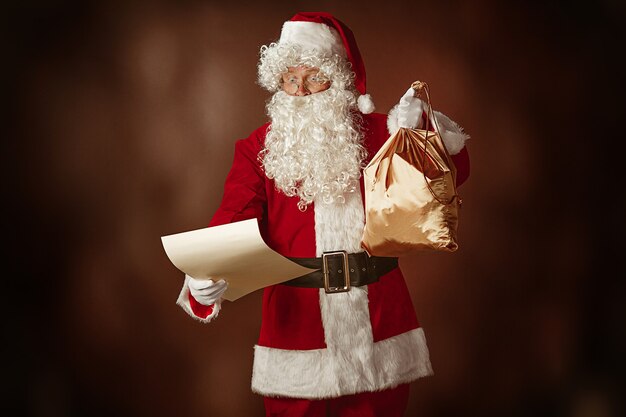 Портрет мужчины в костюме Санта-Клауса - с роскошной белой бородой, шляпой Санта-Клауса и красным костюмом читает письмо на красном фоне студии с подарками