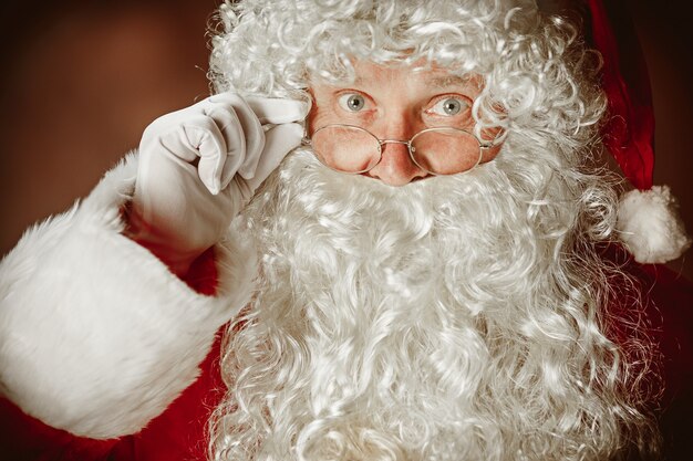 Portrait of Man in Santa Claus Costume - with a Luxurious White Beard, Santa's Hat and a Red Costume at red studio background. The face close up