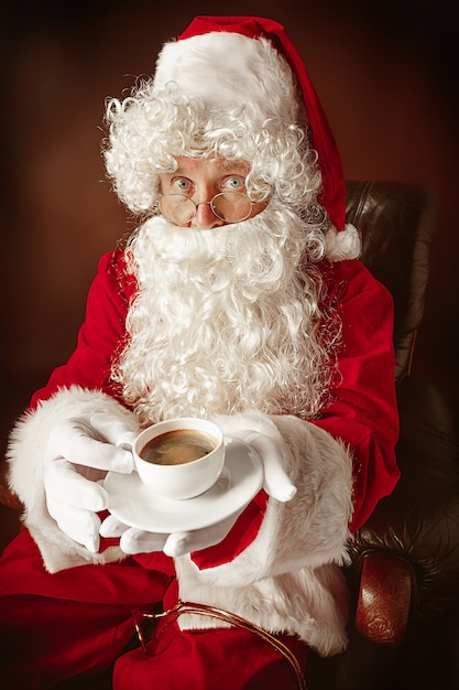 サンタクロースの衣装を着た男の肖像-豪華な白ひげ、サンタさんの帽子、コーヒーのカップが付いている椅子に座っている赤いスタジオの背景に赤い衣装