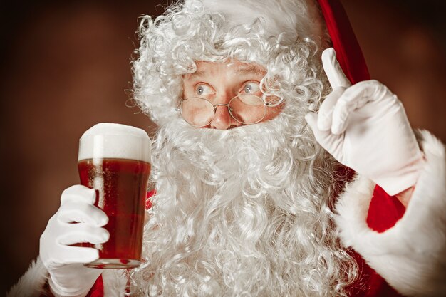 サンタクロースの衣装の男の肖像-豪華な白ひげ、サンタさんの帽子、ビールと赤いスタジオの背景に赤い衣装