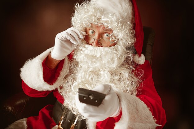 Портрет мужчины в Санта-Клаусе с роскошной белой бородой, шляпой Санта-Клауса и красным костюмом на красном фоне студии