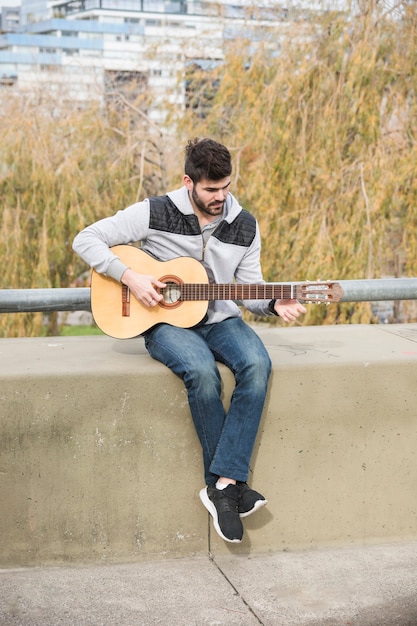 Portrait of a man sitting on retaining wall playing guitar