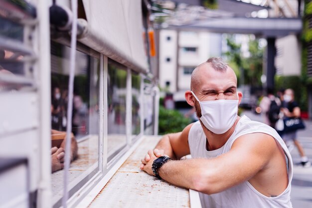 Портрет мужчины в белой медицинской маске на центральной площади города сидит на стуле в кафе-фургоне