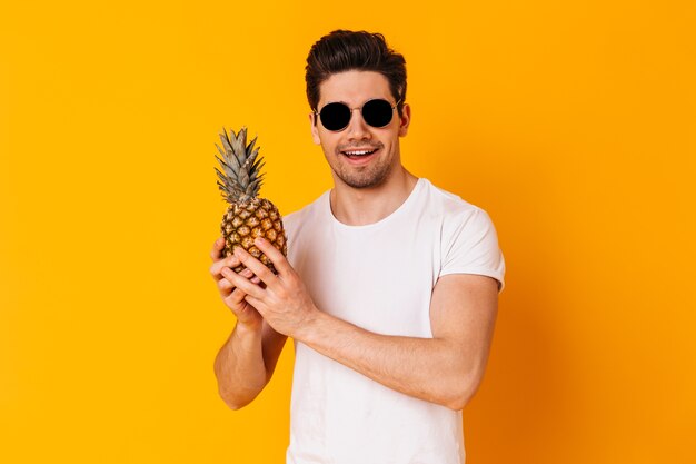 Портрет мужчины в белой футболке и солнцезащитных очках, держа ананас на оранжевом пространстве.