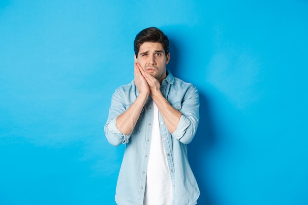 Portrait of man with toothache grimacing from pain and touching cheek, standing against blue background