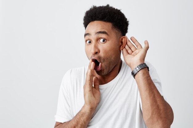 Portrait of mature beautiful black-skinned shocked male with afro hairstyle in casual white t-shirt holding hand near mouth, listening gossips about his boss with surprised expression