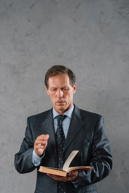 Портрет зрелого бизнесмена, чтение книги, стоя на фоне серый текстурированные