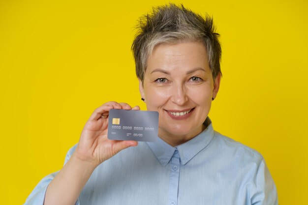 黄色の背景に分離されたオンライン支払いまたはオンラインショッピングを手にクレジットデビットカードを持つ成熟した白髪の女性の肖像オンラインバンキングの概念
