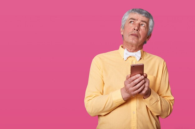 Portrait of mature man having wrinkles and gray hair dressed in yellow shirt and white bow tie, holding smartphone in hands, looks up. Elderly man with mobile phone posing in studio isolates on pink.