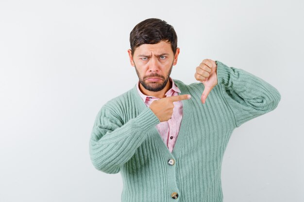 Portrait of mature man pointing right, showing thumb down in shirt, cardigan and looking upset front view