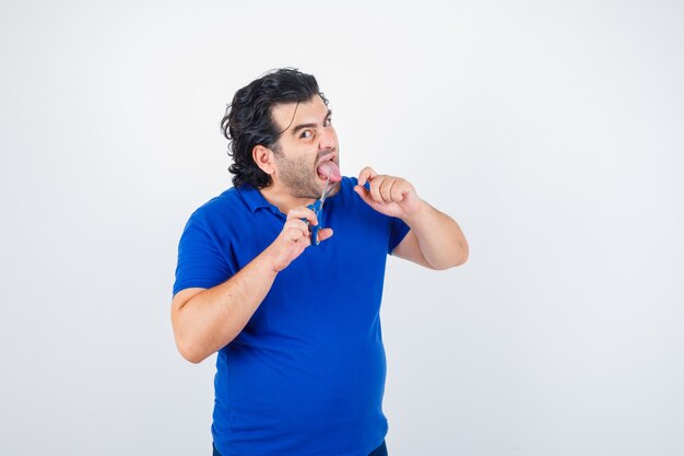Портрет зрелого мужчины в синей футболке, который пытается отрезать себе язык ножницами и выглядит агрессивно, вид спереди